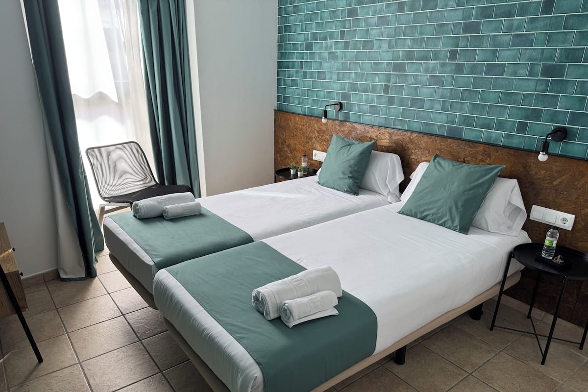 Ein Hotelzimmer mit zwei Betten, Bettwäsche in Türkis und Weiß, einem Holzkopfstück, einem kleinen Tisch mit einer Flasche und einem Stuhl.