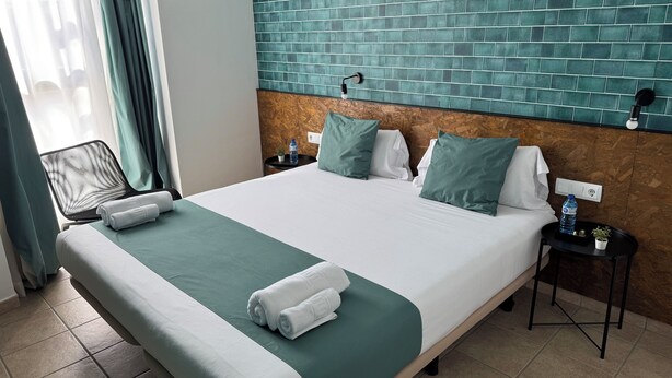 Une chambre d’hôtel avec un grand lit, une tête de lit ornée d’un mur recouvert de carreaux verts, une petite table de chevet avec une bouteille d’eau et une chaise près de la fenêtre.