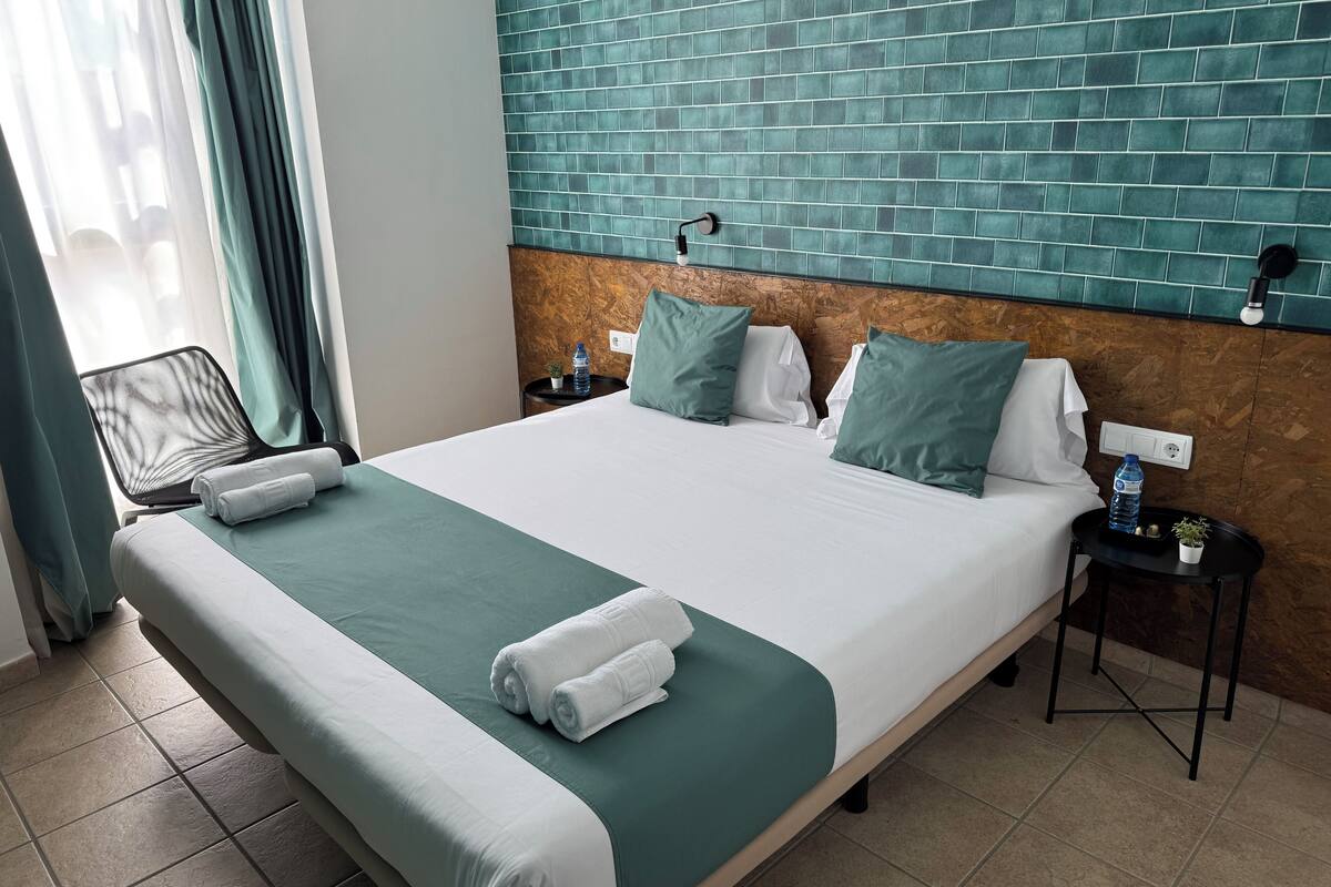 Ein Hotelzimmer mit einem großen Bett, einer Wand mit grünen Fliesen als Kopfteil, einem kleinen Nachttisch mit einer Wasserflasche und einem Sessel in der Nähe des Fensters.