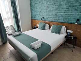 Habitación de hotel con una cama grande, cabecera con pared de azulejos verdes, mesita de noche con botella de agua y una silla cerca de la ventana.