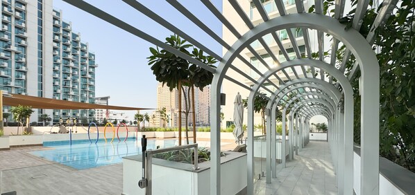 Un espace piscine extérieur moderne, doté d’une barrière en verre, d’une pergola en métal et offrant une vue sur les gratte-ciel en arrière-plan.