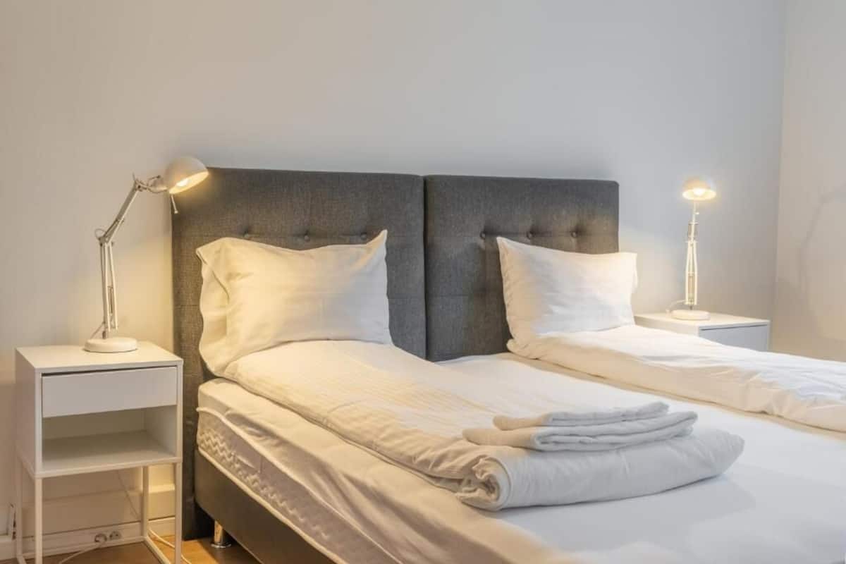 A neatly made bed with white linens and pillows, two bedside tables with lamps, and a grey upholstered headboard.