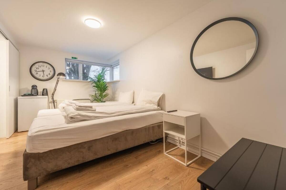 A bedroom with a bed, bedside table, a round mirror, a clock, and a window.