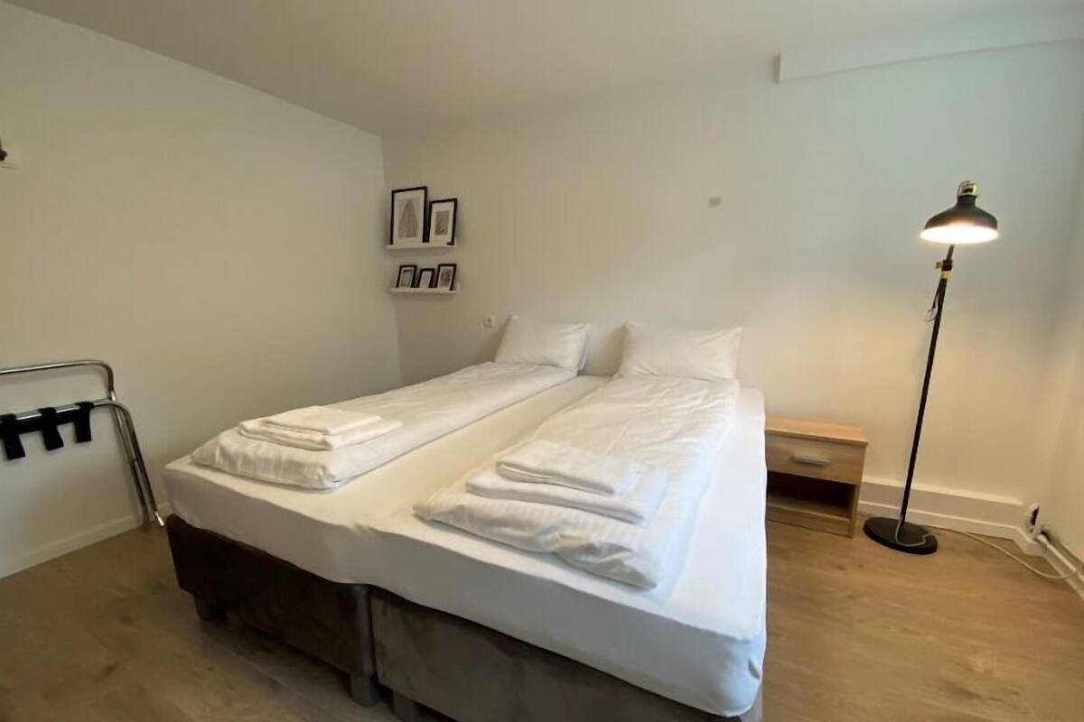 A double bed with white linens, a bedside table, a floor lamp, and a towel rack.