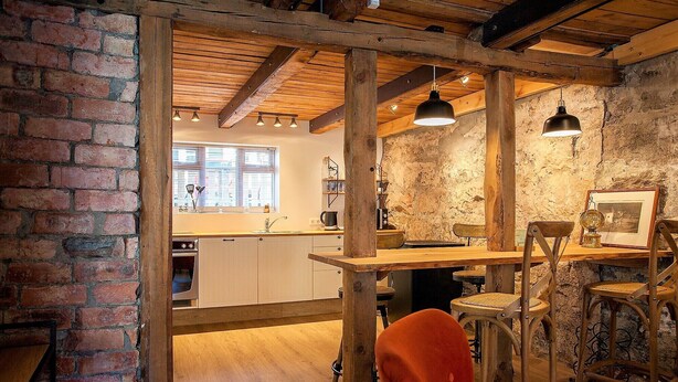 A rustic kitchen with exposed wooden beams, a brick wall, and a wooden dining table with chairs.