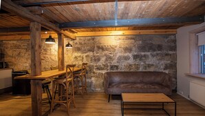 A rustic kitchen with wooden beams, a stone wall, a brown leather sofa, a wooden table, and bar stools.