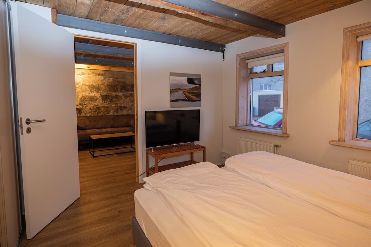 A bedroom with a bed, a television on a wooden stand, a stone wall, and a window with a view of a red car.