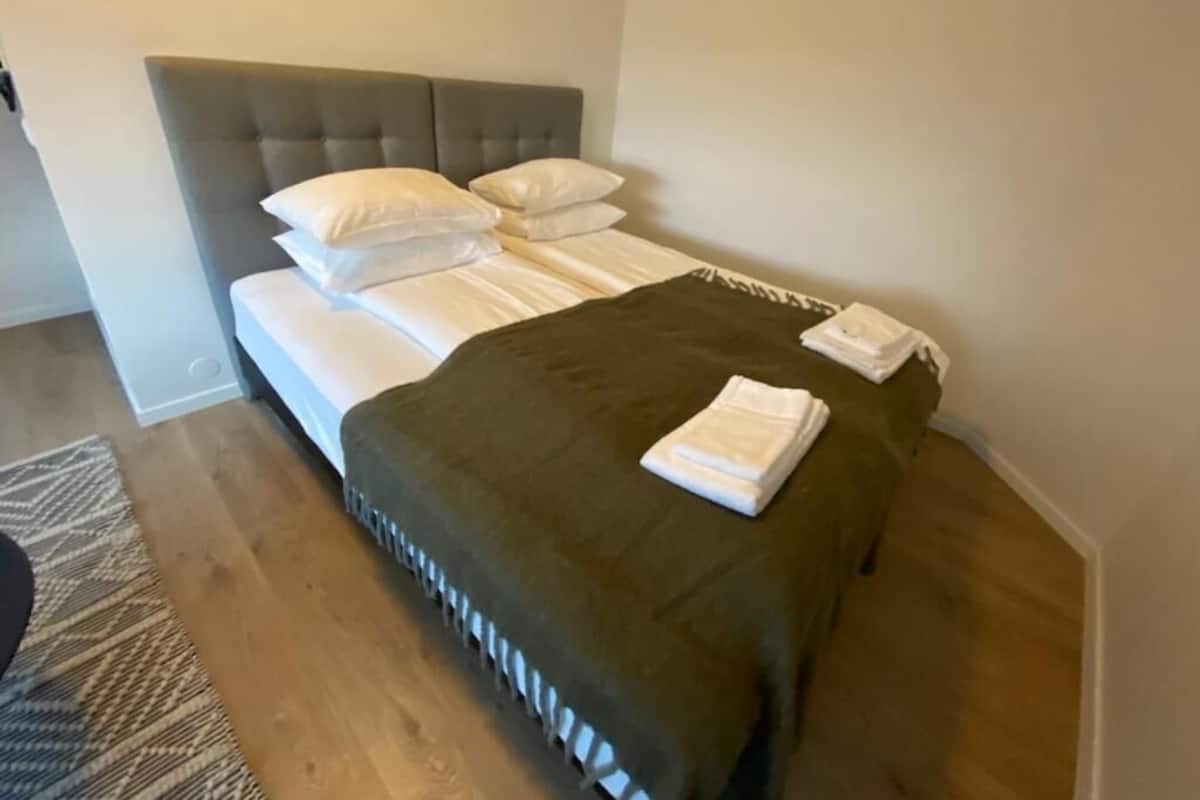 A neatly made bed with white linens and a dark green throw blanket, a grey upholstered headboard, and a white pillow on the left side.