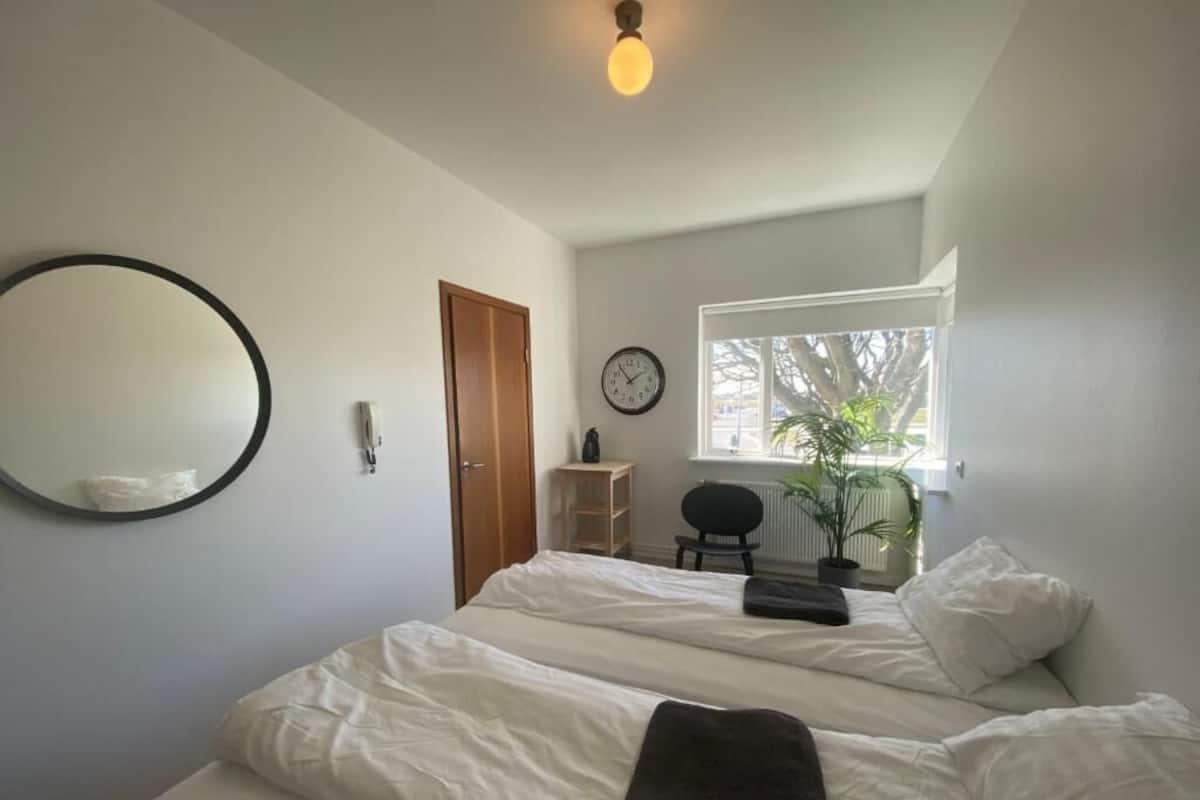 A bedroom with two beds, a round mirror, a clock, a window with a view of trees, and a ceiling light.