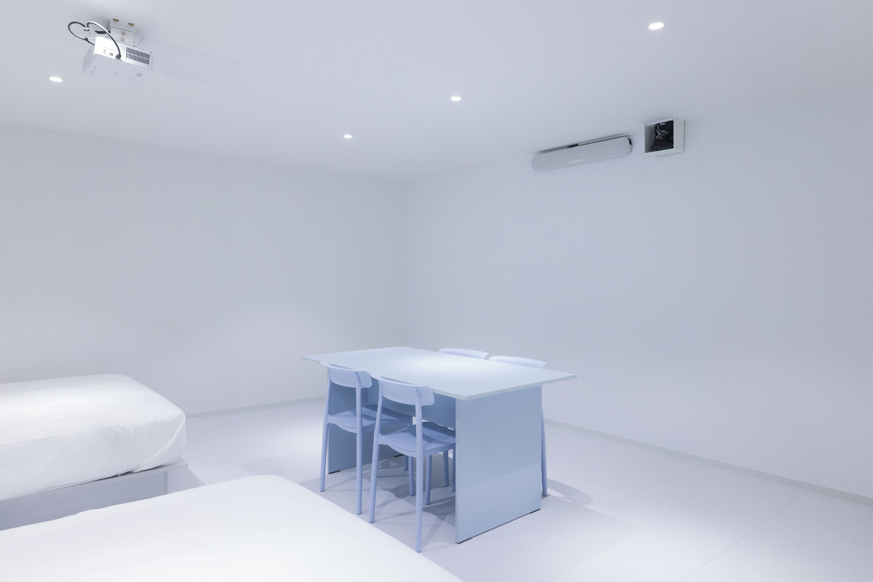A minimalist room with a white bed, a small table with chairs, and a ceiling projector.