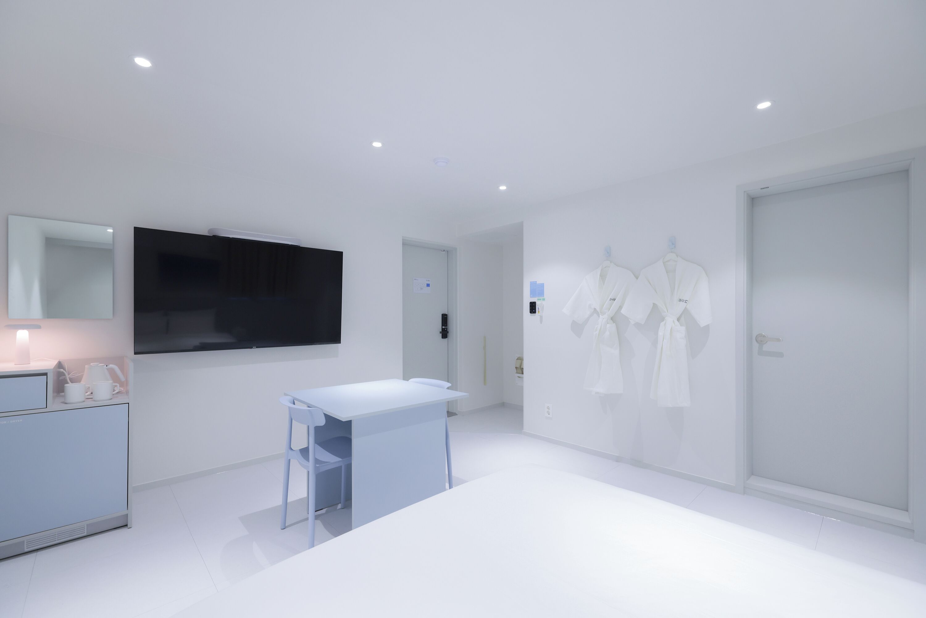 Una habitación moderna y minimalista con una televisión de pantalla plana montada en la pared, un escritorio pequeño con una silla y un armario con ropa colgada.