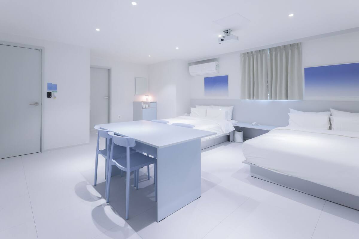 A modern hotel room with a white bed, a desk with a chair, a nightstand, and a ceiling-mounted projector.