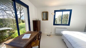 A bedroom with a bed, a wooden desk with a chair, a radiator, a floor lamp, and a window with a view of greenery.