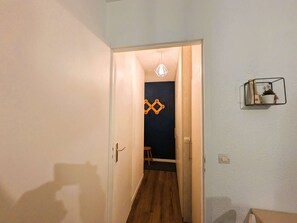 A hallway with a wooden floor, a door leading to a room with a dark wall and a geometric decoration, and a small shelf with a plant and a star decoration. - Modern 1-bedroom apartment with pool, near skiing and village (Arêches-Beaufort)