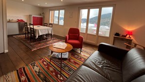 A living room with a sofa, a red chair, a dining table with chairs, a kitchen area, and large windows with a view of mountains. - Charming 2-room apartment in Villard de Lans center (Villard-de-Lans)