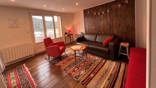 A living room with a wooden floor, a red armchair, a black leather sofa, a small table, and a colorful rug. - Charming 2-room apartment in Villard de Lans center (Villard-de-Lans)