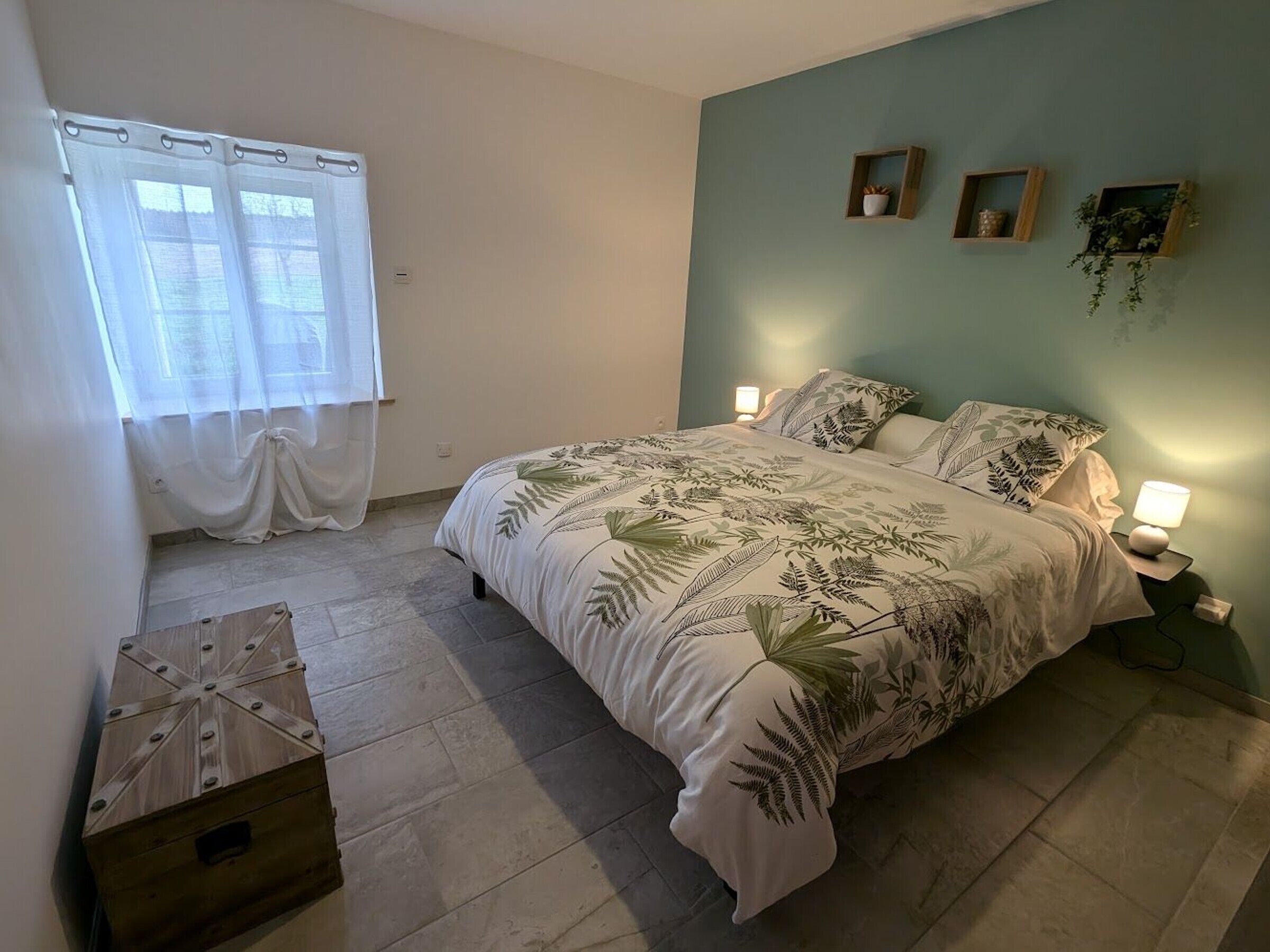A bedroom with a bed, a window with curtains, a bedside table with a lamp, and a small wooden box.
