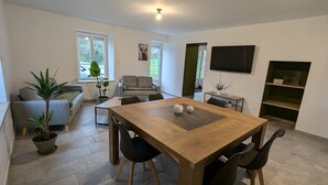 Un salon moderne avec une table à manger en bois, deux chaises noires, un canapé gris, un téléviseur fixé au mur et une plante en pot.