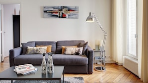 A modern living room with a grey sofa, a coffee table with books and a vase, a floor lamp, and a painting on the wall. - Beauregard Design Résidence (Paris)