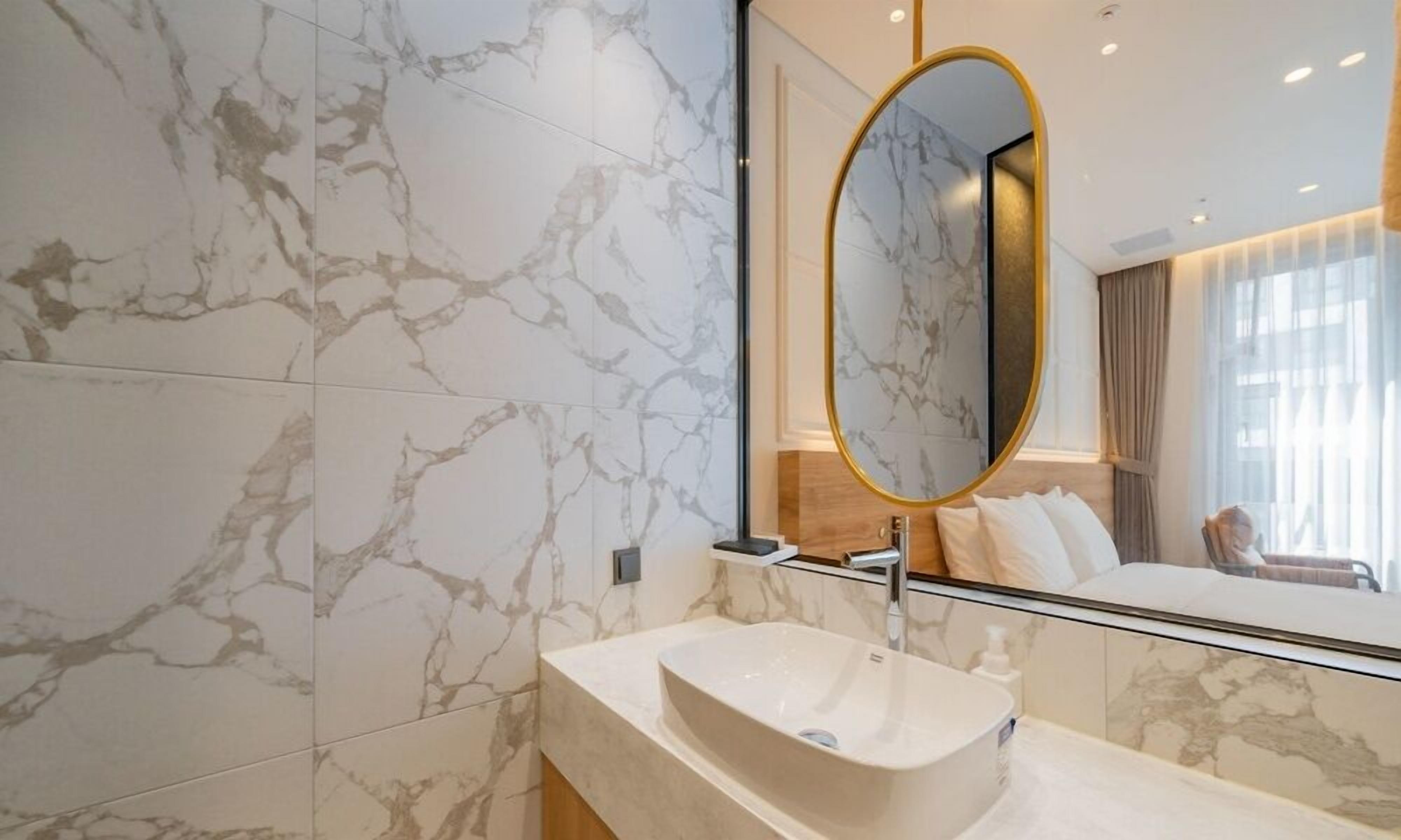 A modern bathroom with marble tiles, a freestanding bathtub, and a large oval mirror with a gold frame.