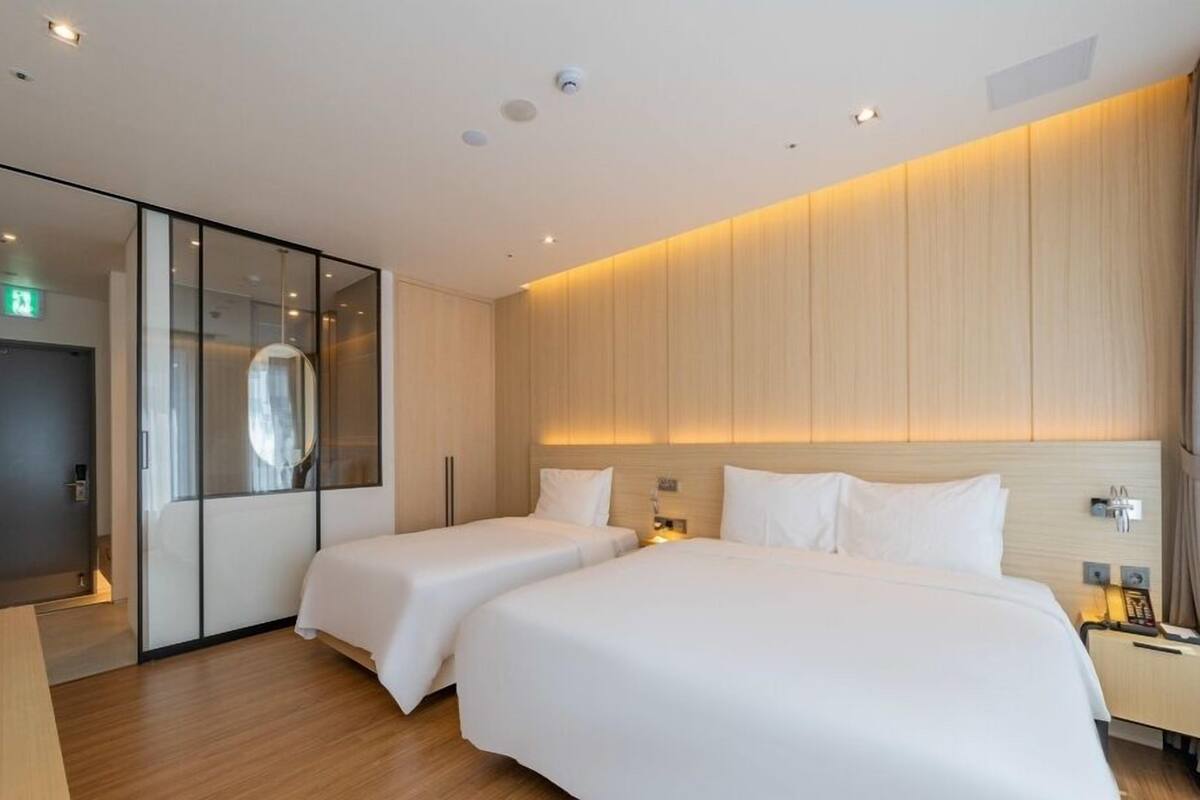 A modern hotel room with two beds, a large mirror, a wardrobe, and a bedside table with a lamp.