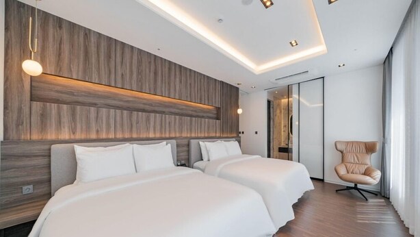 A modern hotel room with two beds, a wooden accent wall, recessed ceiling lighting, and a seating area with a chair. - THE HOUND HOTEL BANDALISLAND 2 (Ansan)