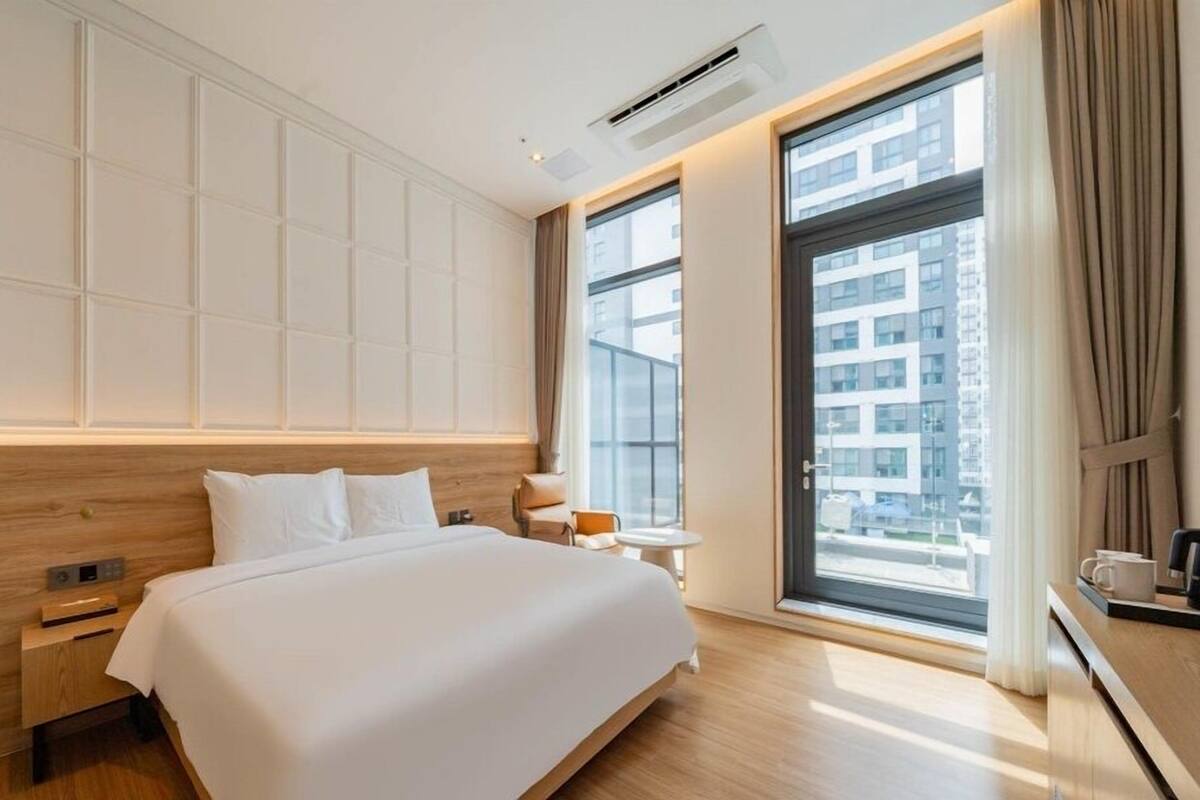 A modern hotel room with a large bed, a small bedside table, a nightstand, a window with a city view, and a built-in air conditioning unit.