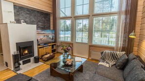 A cozy living room with a fireplace, a sofa, a coffee table, and large windows offering a view of trees. - Rukan tunturituuli by Interhome (Kuusamo)
