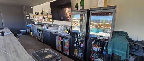 A bar area with multiple refrigerators stocked with beverages, a counter with glasses and bottles, and a view of a pool area through the glass.