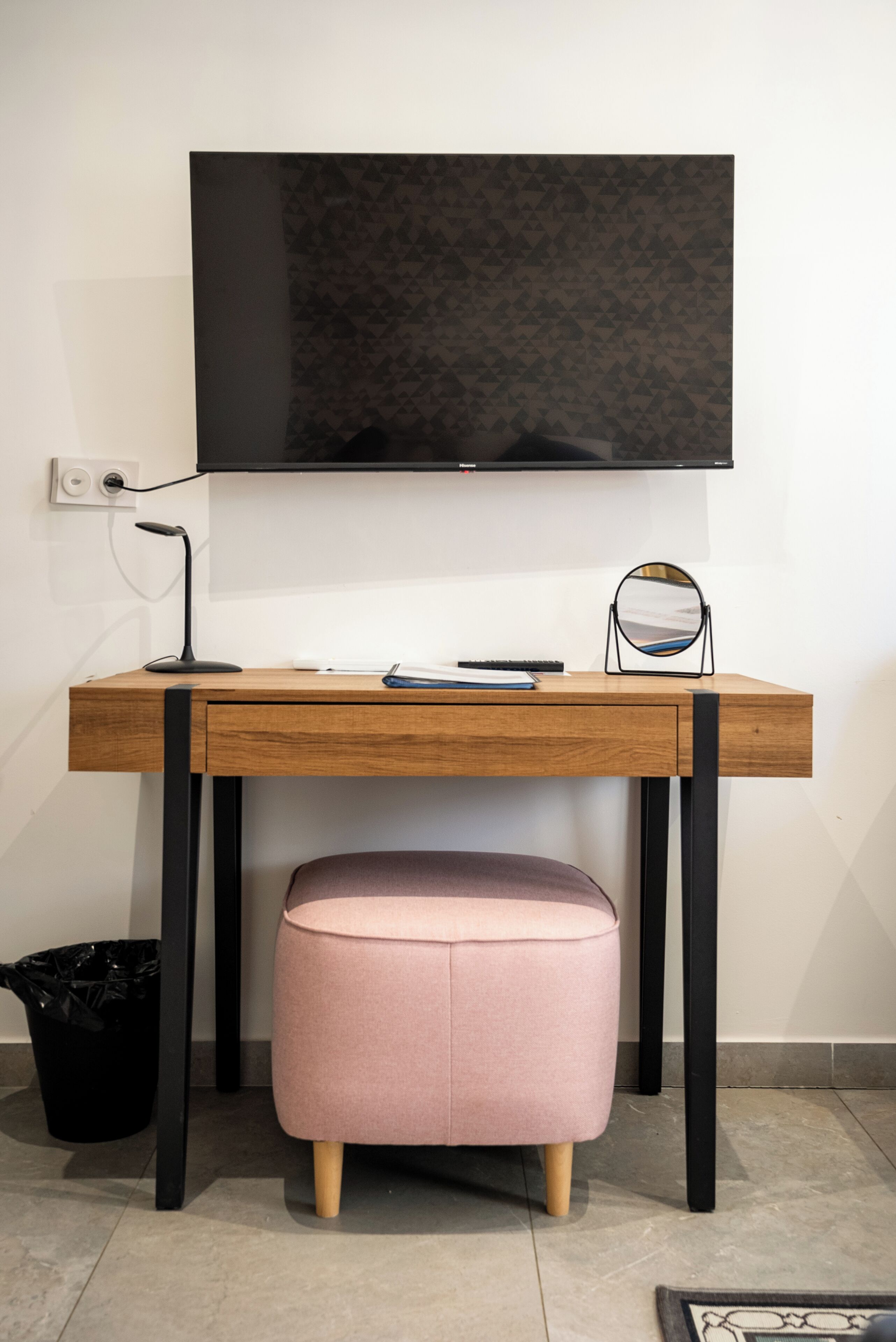 Un escritorio de madera con un televisor de pantalla plana, una lámpara de escritorio, un espejo y un otoman rosa.