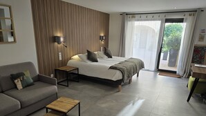 A modern bedroom with a bed, a sofa, a small wooden table, and a view of the outdoors through a large window. - Les Loges du Luberon (Cabrieres-d'Avignon)