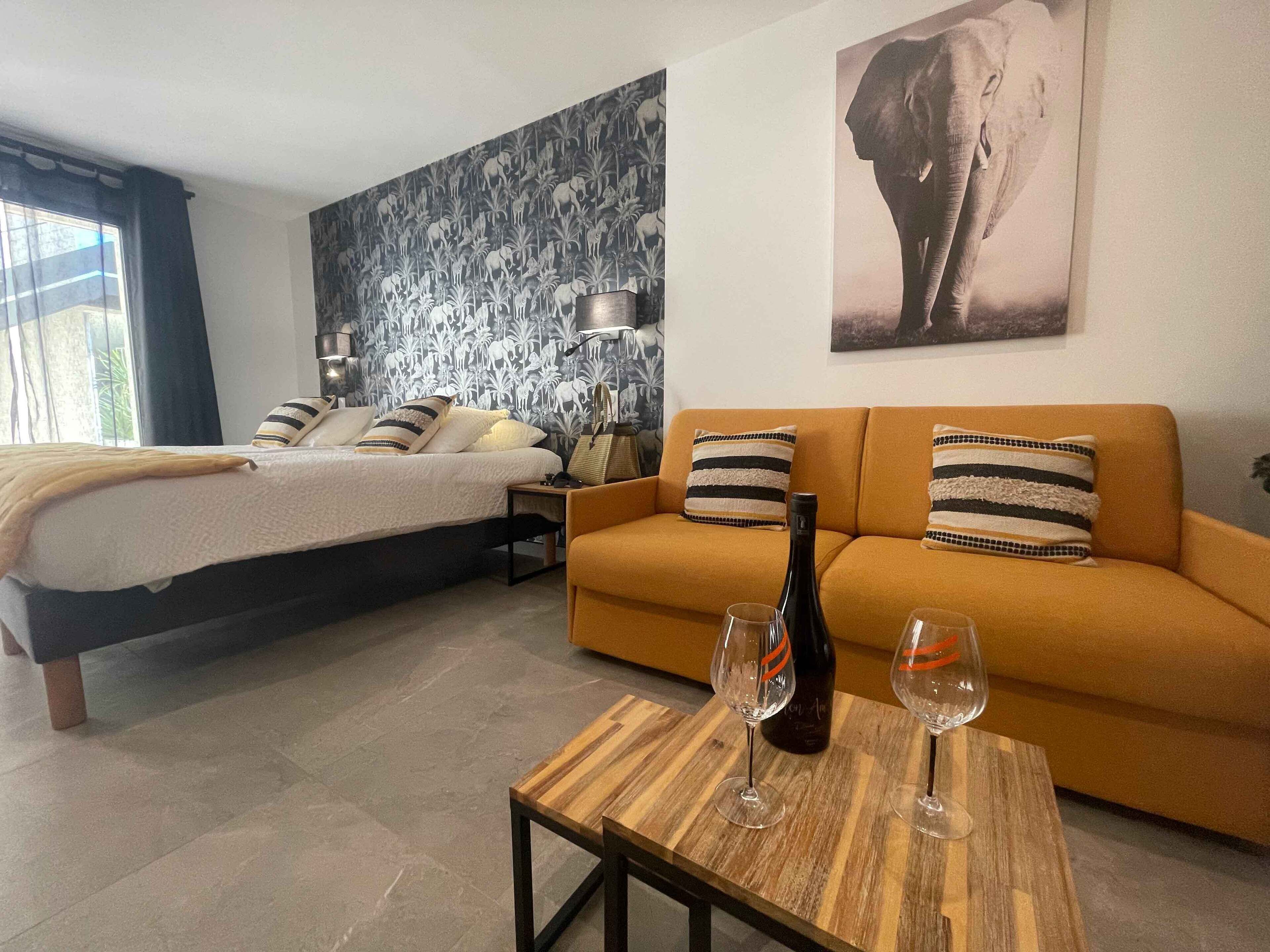 A bedroom with a bed, a sofa, a table with wine glasses, and a wall with a giraffe and elephant painting.