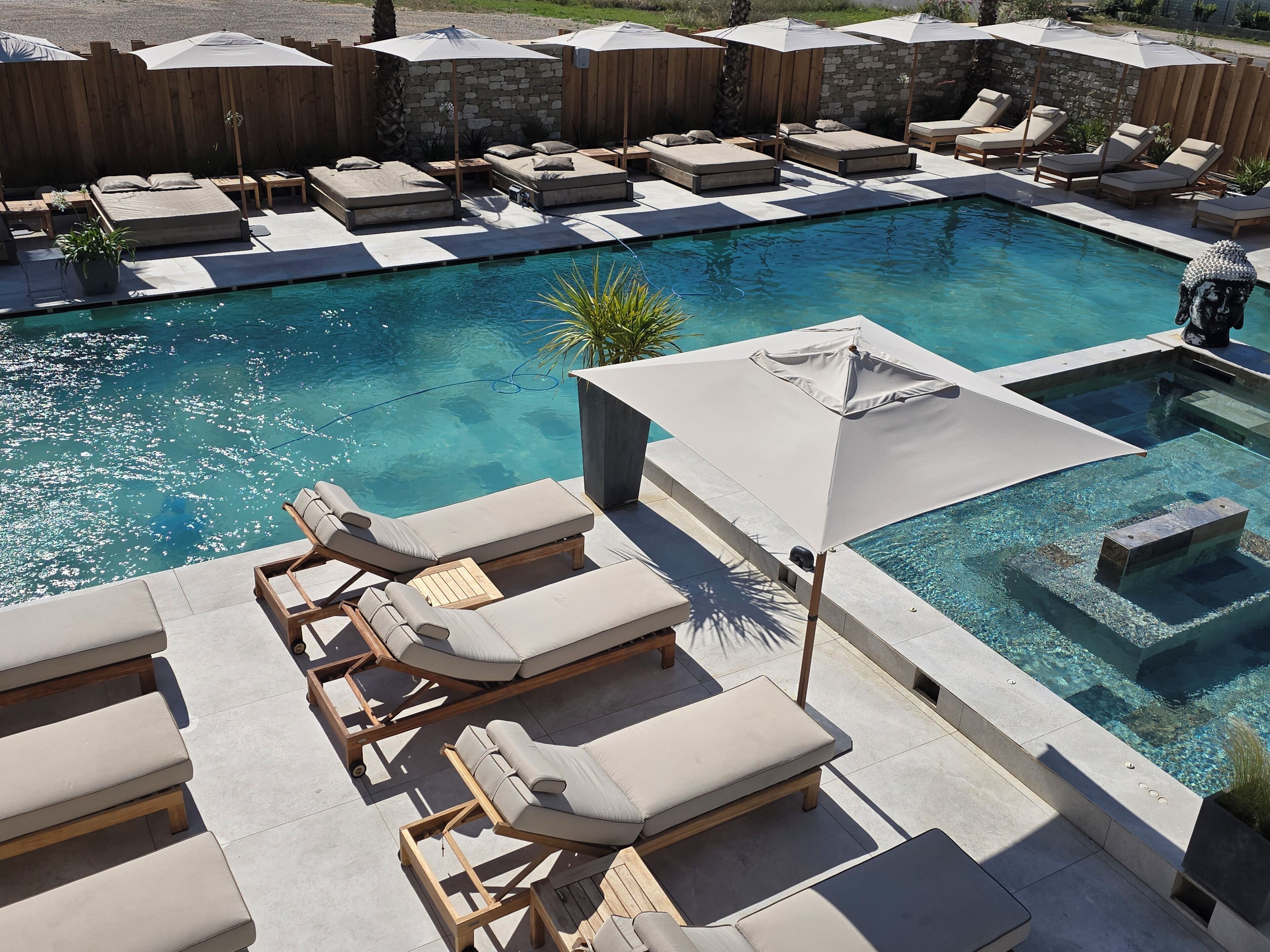 A poolside area with lounge chairs and umbrellas, a stone wall, and a wooden fence.
