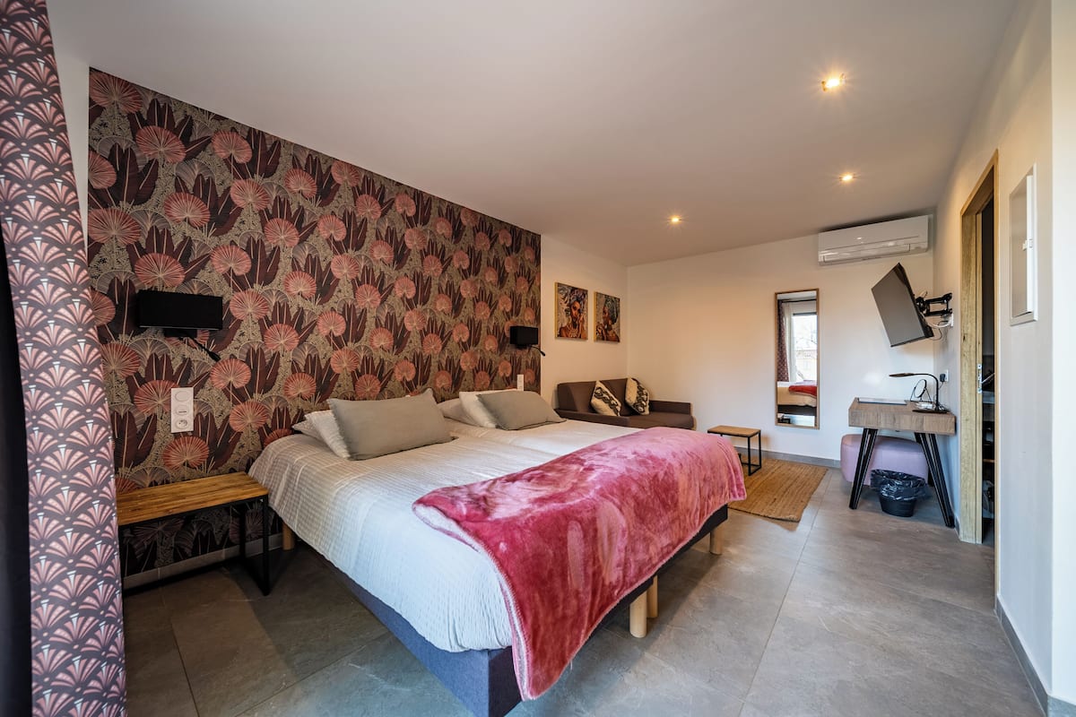Une chambre avec un grand lit, un banc en bois, une couverture rouge, un téléviseur fixé au mur et un miroir.