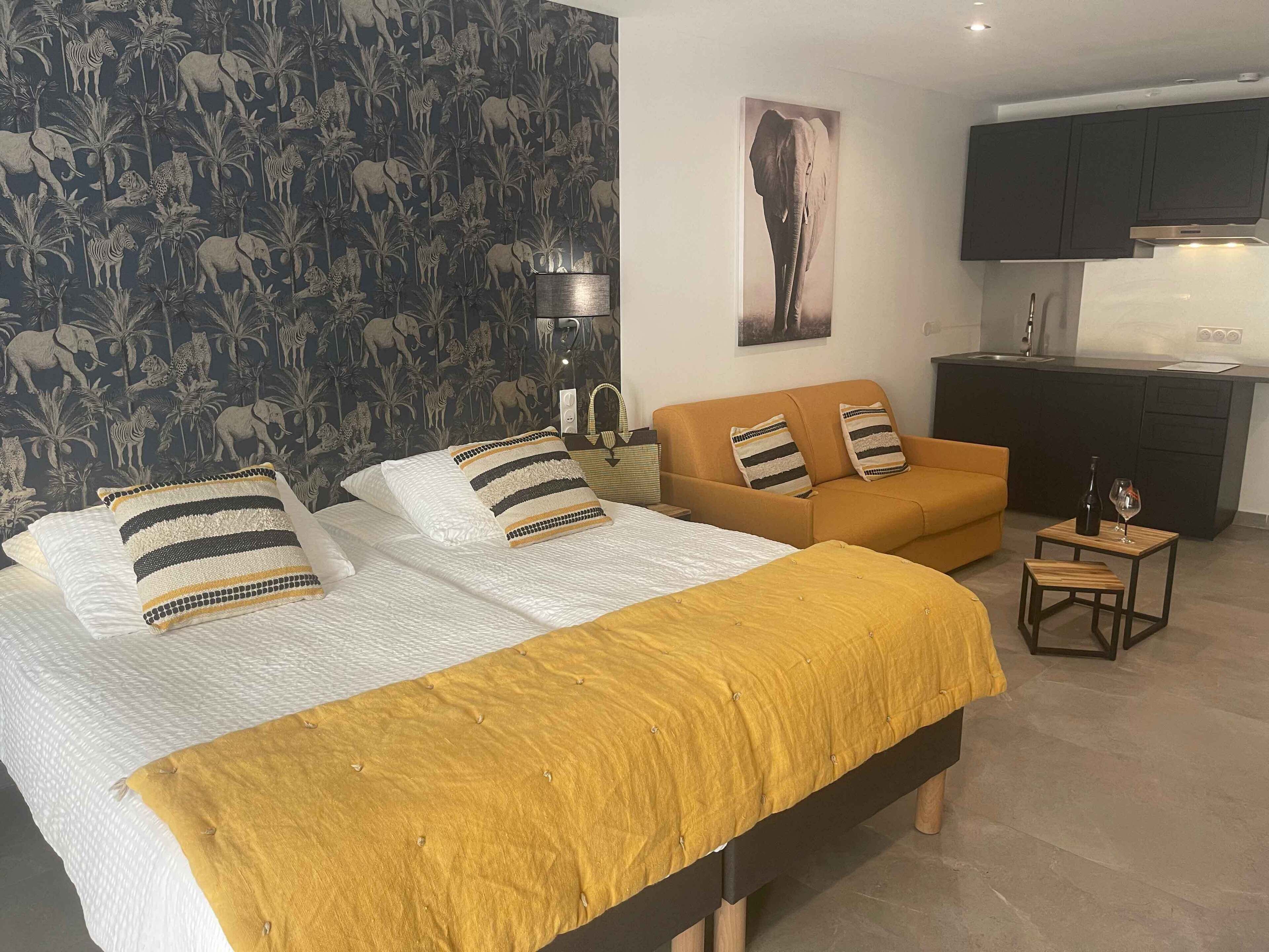 Un dormitorio con dos camas, un sofá, una mesita con una botella y un vaso, y una zona de cocina al fondo.