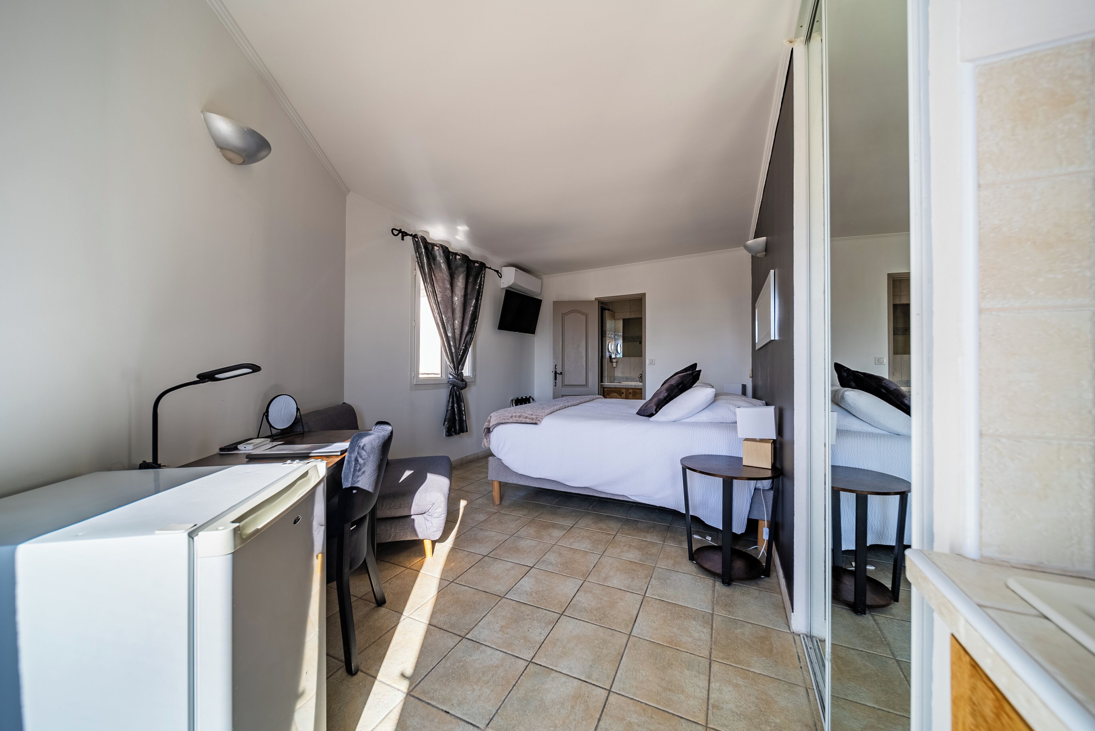 Quarto de hotel compacto com cama, escrivaninha, cadeira, mesa pequena e televisão.