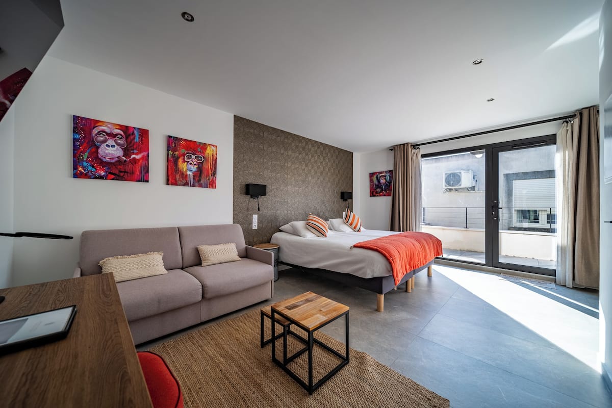 Une chambre moderne avec un lit, un canapé et une petite table en bois. Deux tableaux sont accrochés au mur, il y a une fenêtre avec des rideaux et on peut voir un appareil de climatisation à travers la fenêtre.