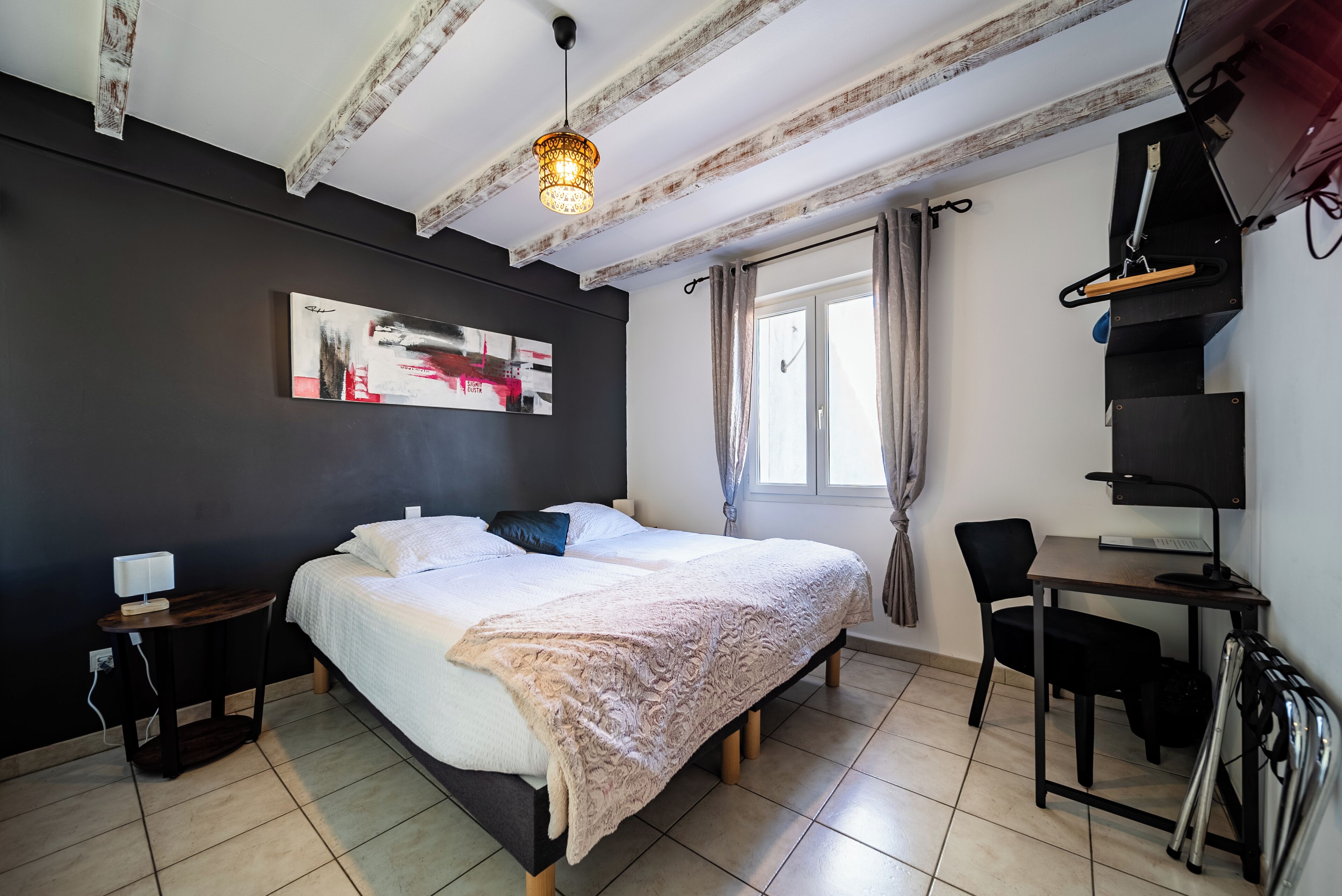 A bedroom with a bed, bedside table, desk, chair, and a hanging light fixture.