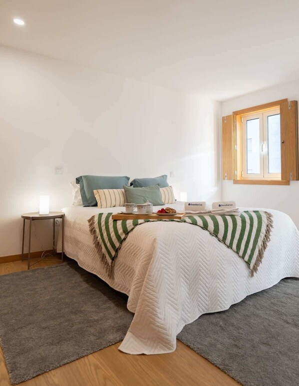 Un dormitorio con cama, almohadas, una mesita de noche, una bandeja con alimentos para el desayuno y una ventana con contraventanas de madera.
