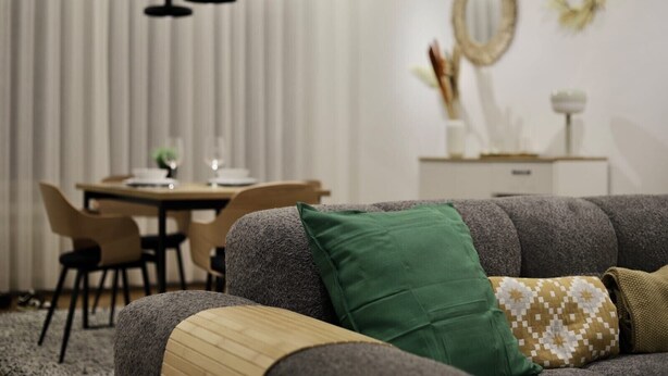 A living room with a grey sofa, a green pillow, and a patterned throw. There is a dining table with chairs in the background. - Appartement Chic et Confortable (Liege)