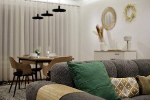 A living room with a grey sofa, a green pillow, and a patterned throw. There is a dining table with chairs in the background. - Appartement Chic et Confortable (Liege)