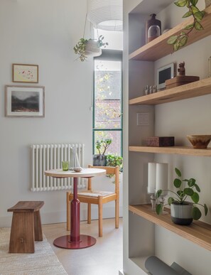 A modern room with a round wooden table, a chair, a radiator, a window with a view of a brick building, and shelves with various items. - Mason & Fifth, Bermondsey (London)
