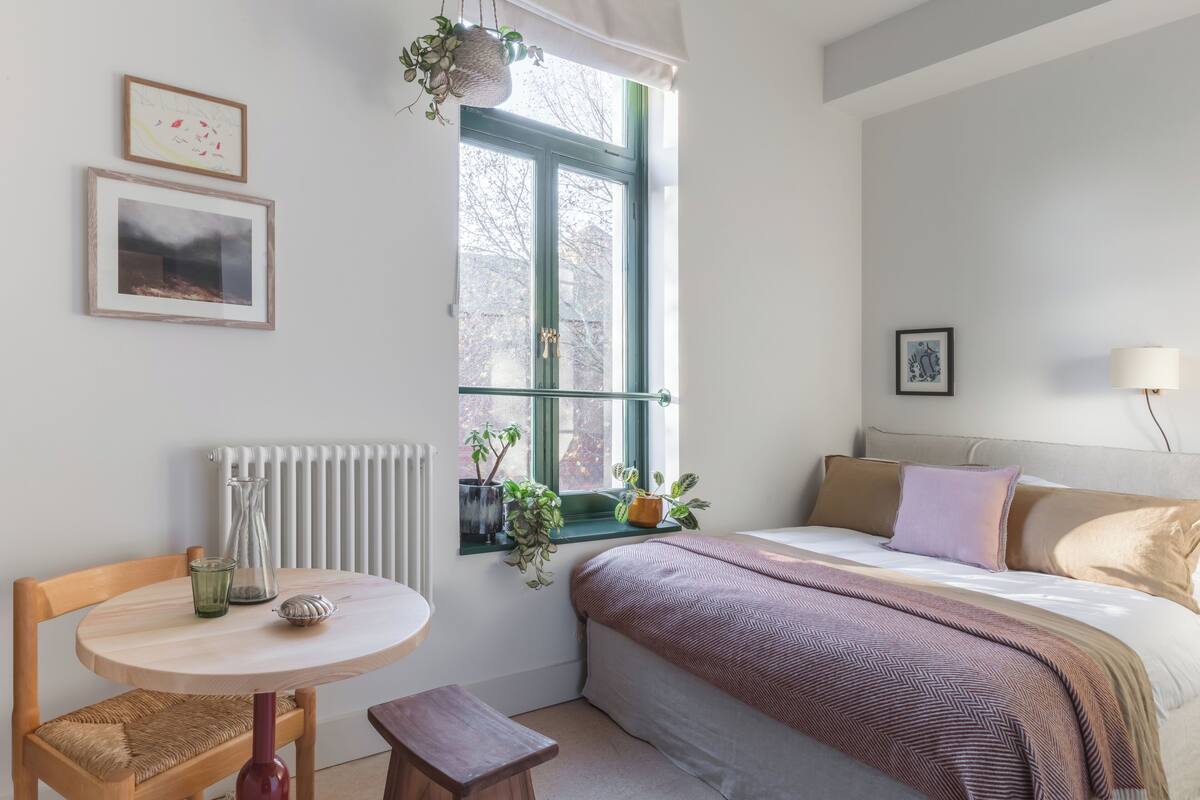 Ein Schlafzimmer mit Bett, Nachttisch, Stuhl und einem Fenster mit Pflanzen.