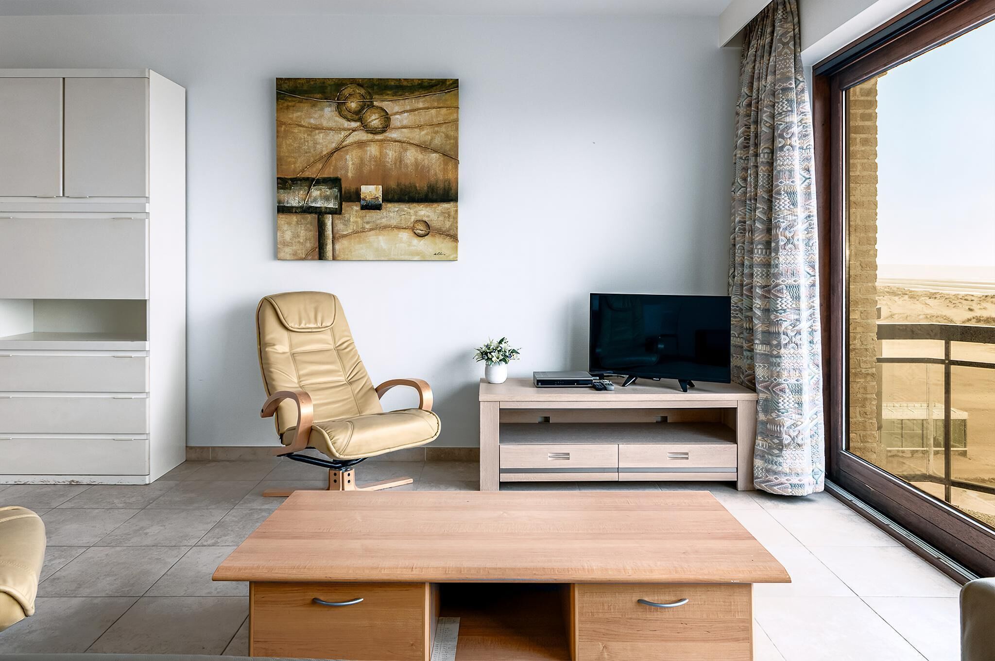 Ein modernes Wohnzimmer mit Couchtisch aus Holz, einem beigefarbenen Ledersessel, einem Fernseher auf Holzfuß und einem großen Fenster mit Blick auf die Wüste.