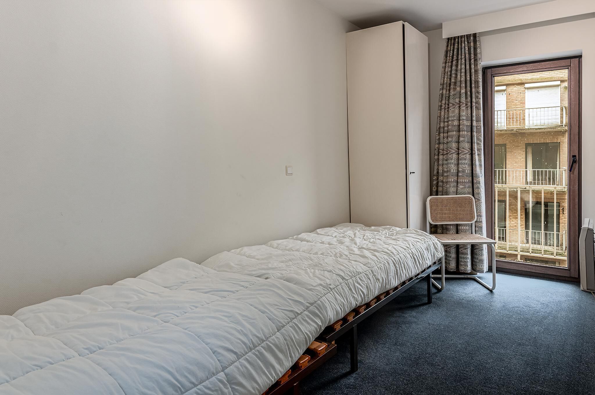 Ein Zimmer mit einem Einzelbett, einem Holzrahmen, einem Stuhl und Blick auf einen Balkon durch ein Fenster mit gemusterten Vorhängen.