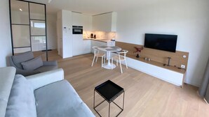 A modern living room with a grey sofa, a coffee table, and a dining area with a white table and chairs. A kitchen with white cabinets and a built-in oven is visible in the background. - 1 bedroom accommodation in Nieuwpoort (Nieuwpoort)