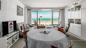 Een moderne woonkamer met een grote televisie, een eethoek met een gestreepte tafelkleed, en uitzicht op het strand door grote ramen.