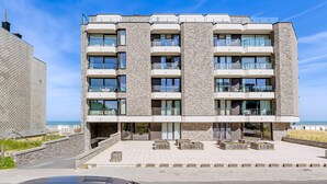 Een hotel met meerdere verdiepingen, modern vormgegeven, met balkons, grote ramen en een verhard buitengebied met zitplaatsen.