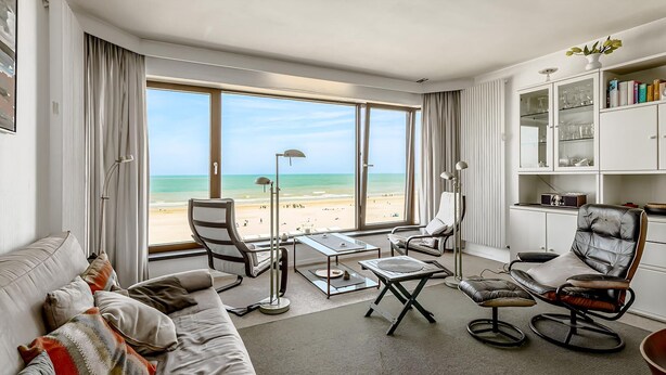 Een moderne hotelkamer met een groot raam met uitzicht op het strand, een leren fauteuil, een bank en een kleine tafel.