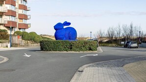 A blue sculpture of a rabbit in a landscaped area with a road and a building in the background.
